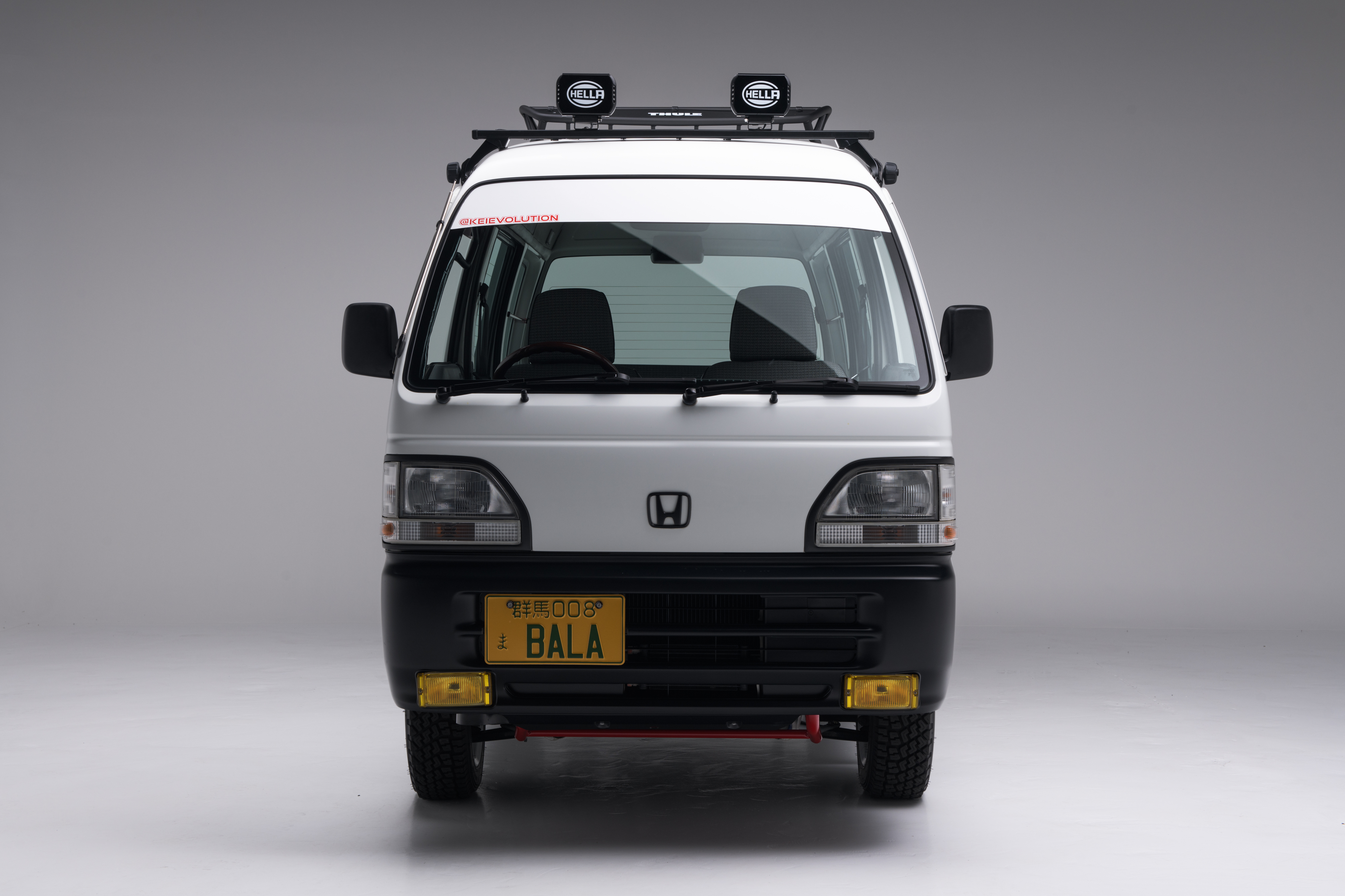 White Honda Acty van with a roof rack and Hella rally lights, front view on a clean studio background